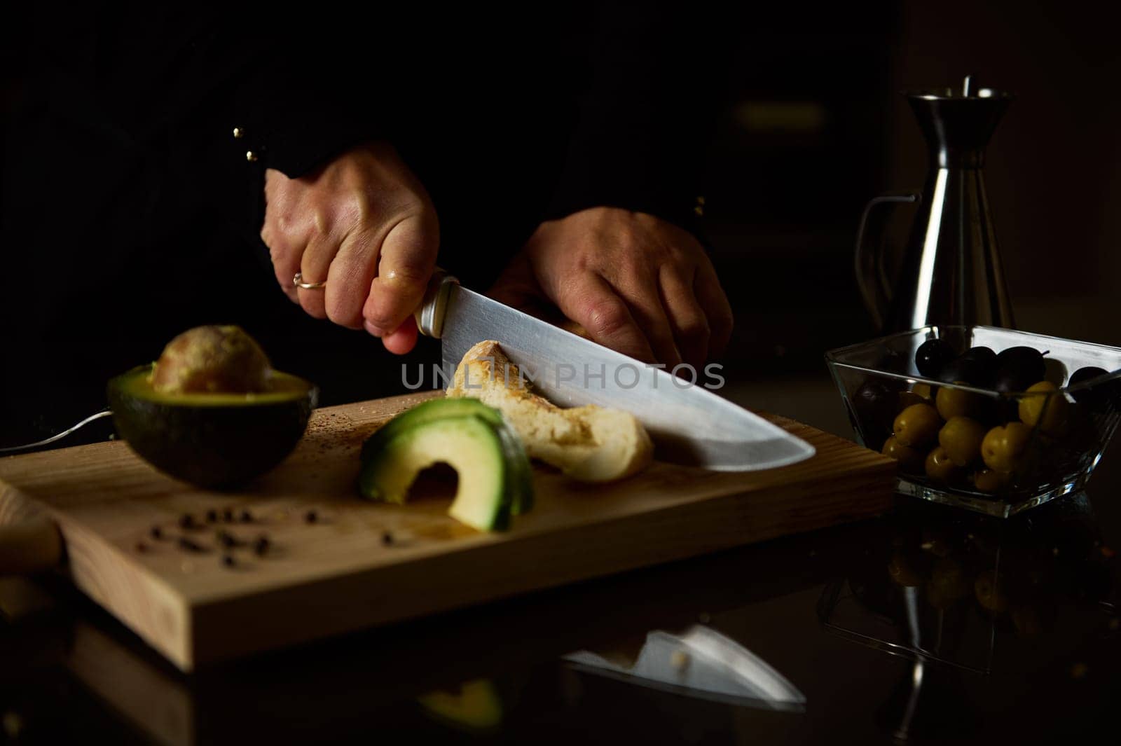 A pair of hands slice bread on a wooden board, surrounded by fresh avocado. The scene captures culinary preparation with a focus on healthy ingredients and home cooking.