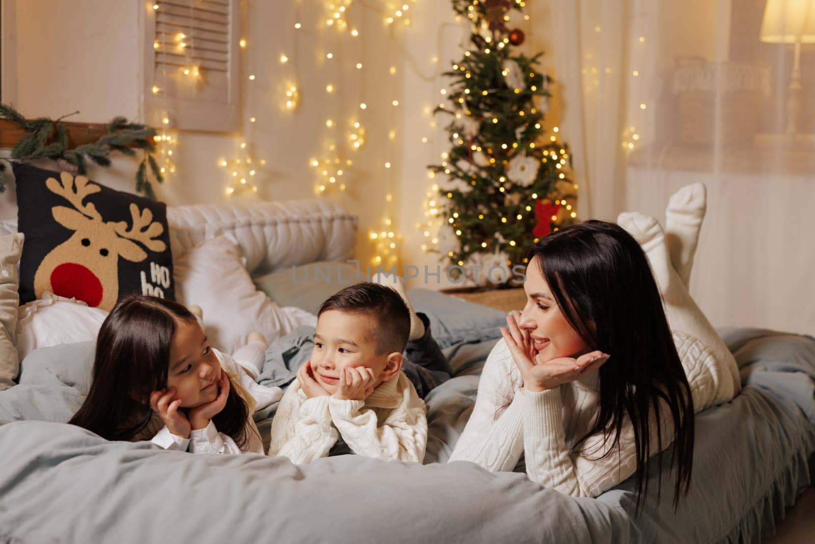 mother and two children lying on bed on christmas happy evening in cozy room decorated with garlands,