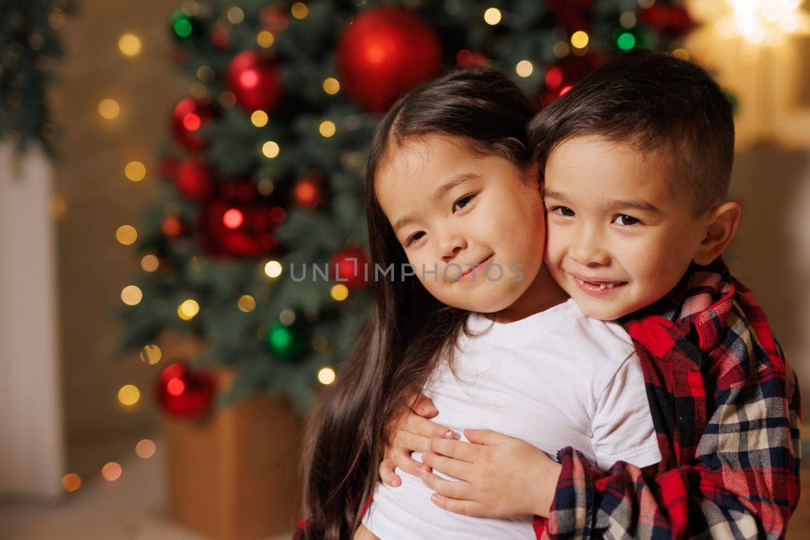 happy asian children hugging against christmas tree background in cozy room decorated with garlands,