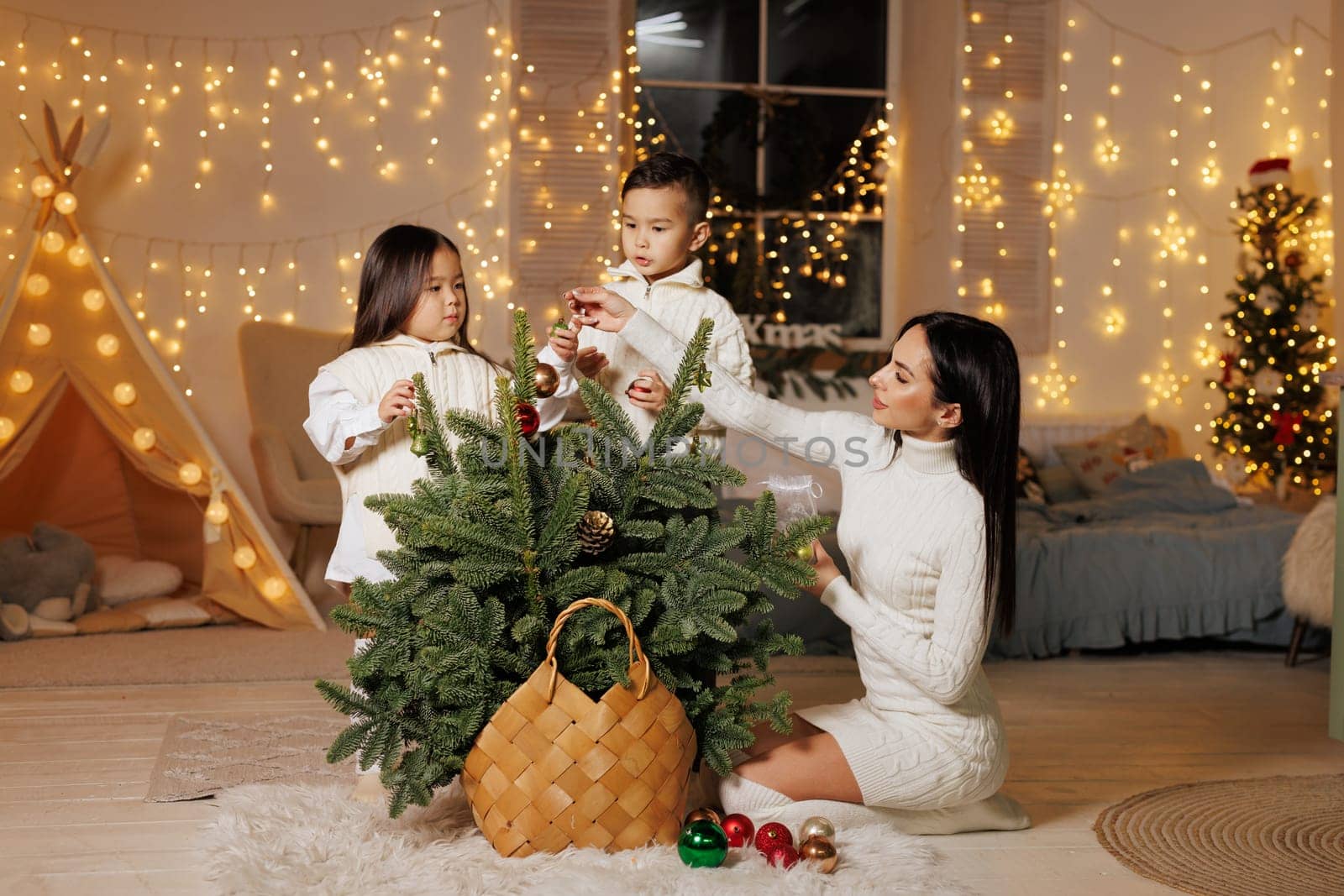 mother and two children decorate a small Christmas tree on Christmas evening in a cozy room decorated with New Year garlands, happy family at Christmas,