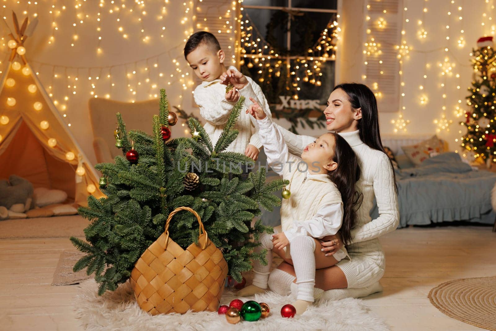A mother and her two children decorating a small Christmas tree in a cozy, festive room adorned with fairy lights. The children are playfully reaching for ornaments, while the mother watches with a smile. The atmosphere is warm and joyful,