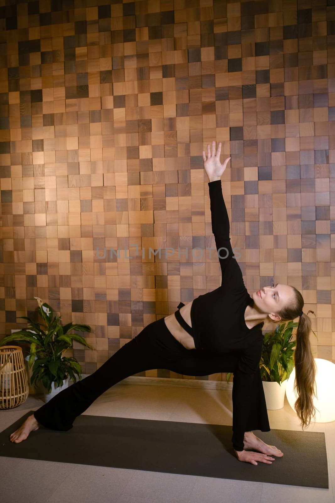 Athletic girl in black sportswear doing yoga in fitness room among indoor green plants.