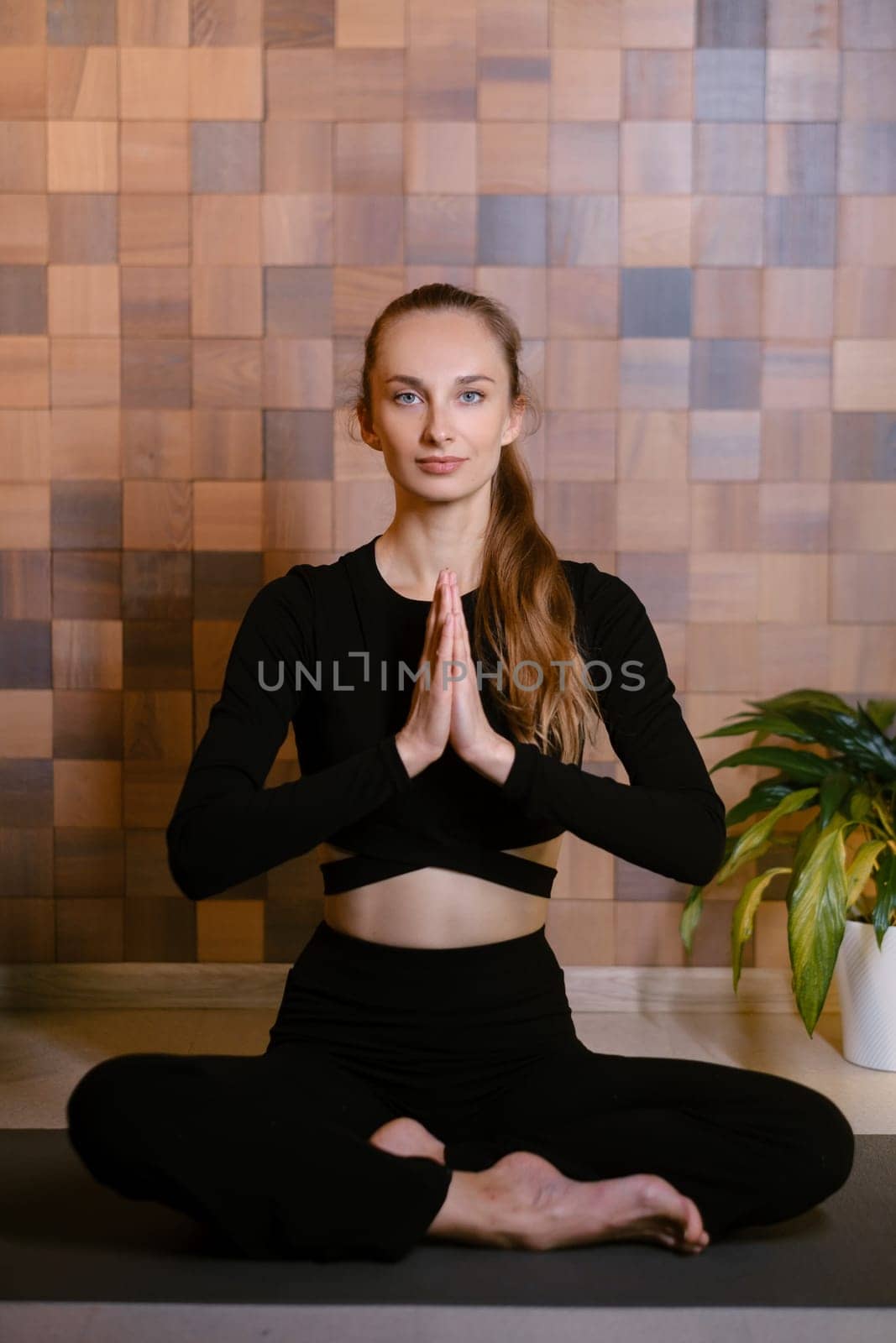 An athletic girl in black sportswear sits in lotus pose at a yoga class in a fitness room among green indoor plants.