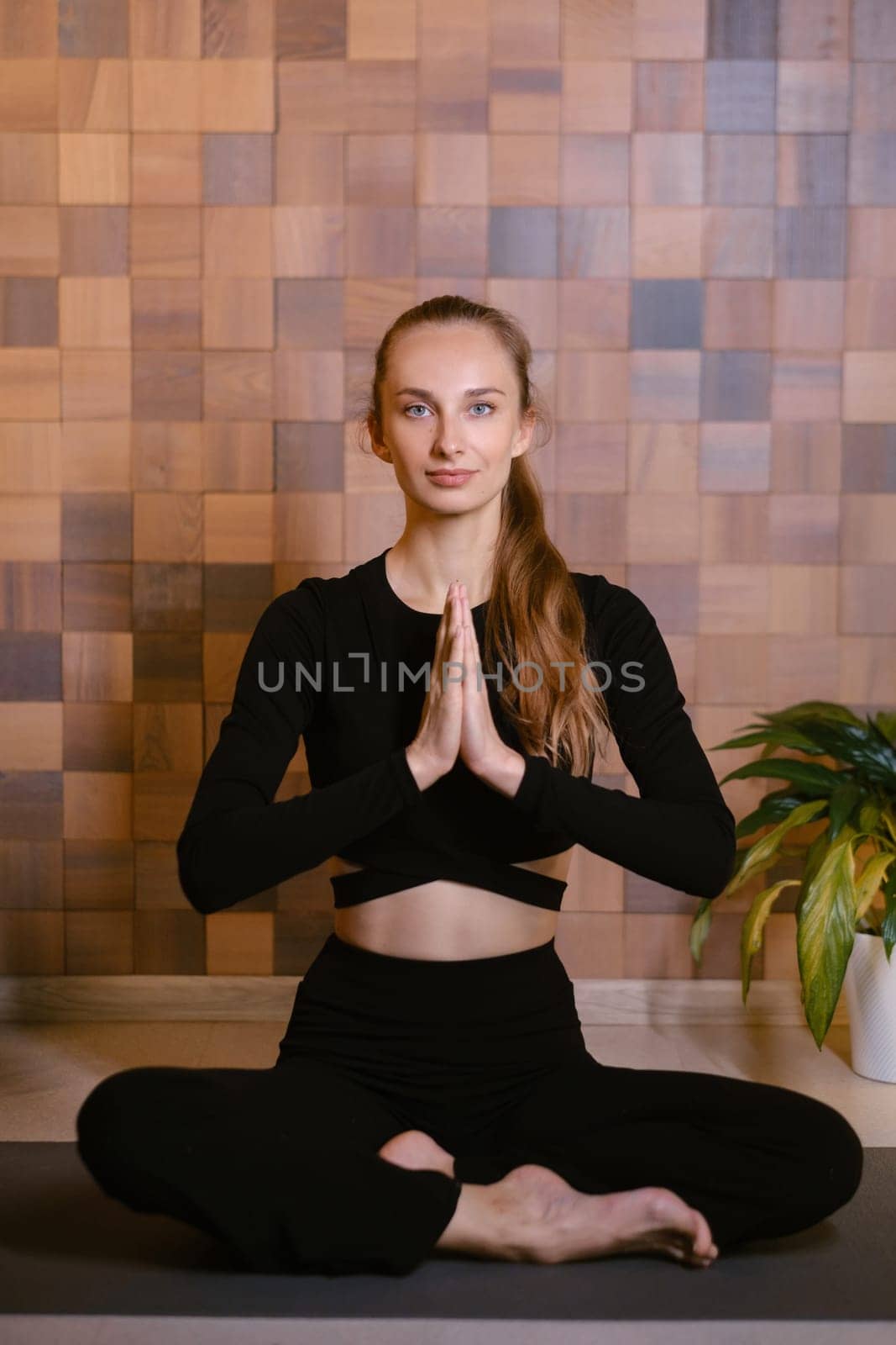 An athletic girl in black sportswear sits in lotus pose at a yoga class in a fitness room among green indoor plants.