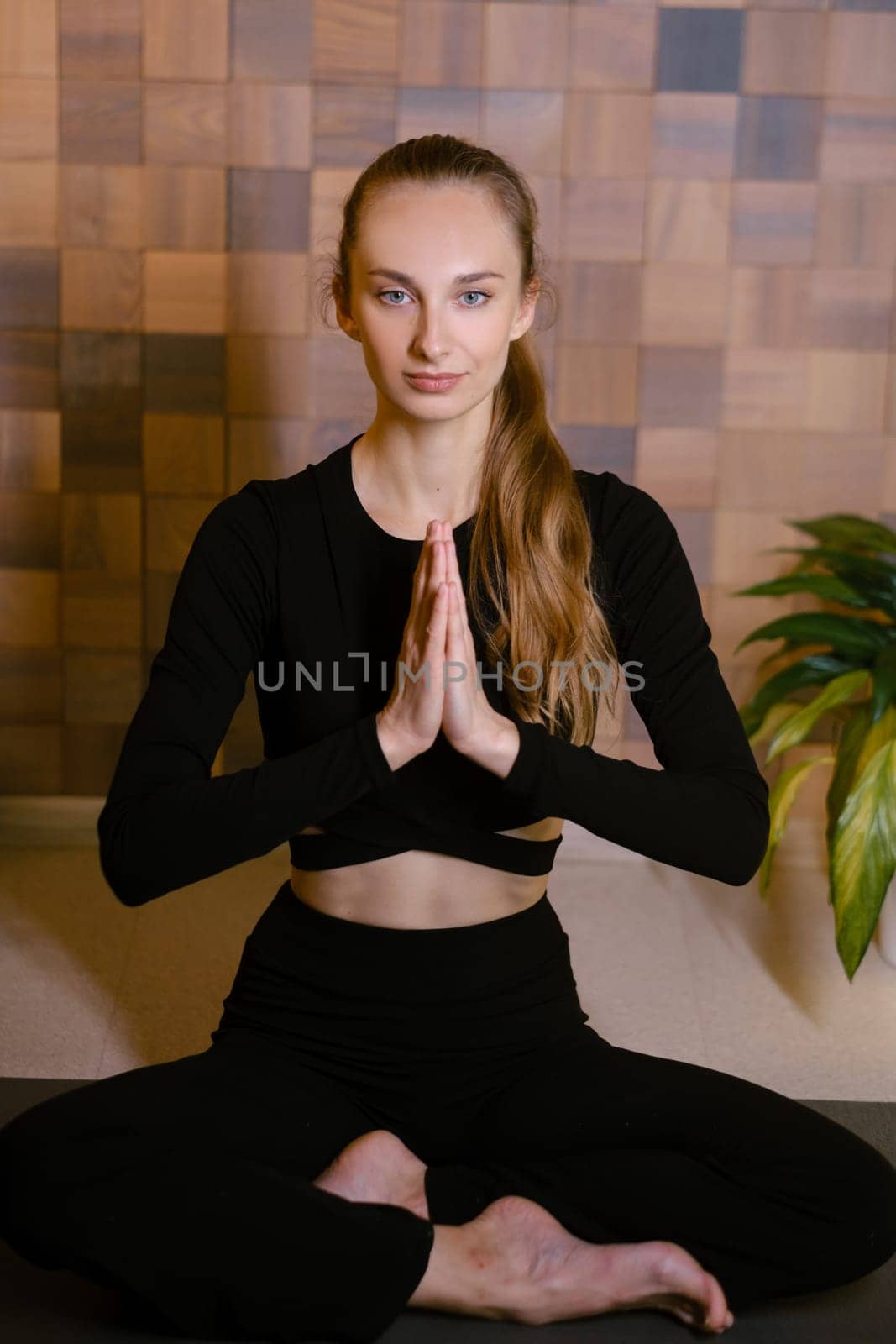 An athletic girl in black sportswear sits in lotus pose at a yoga class in a fitness room among green indoor plants.