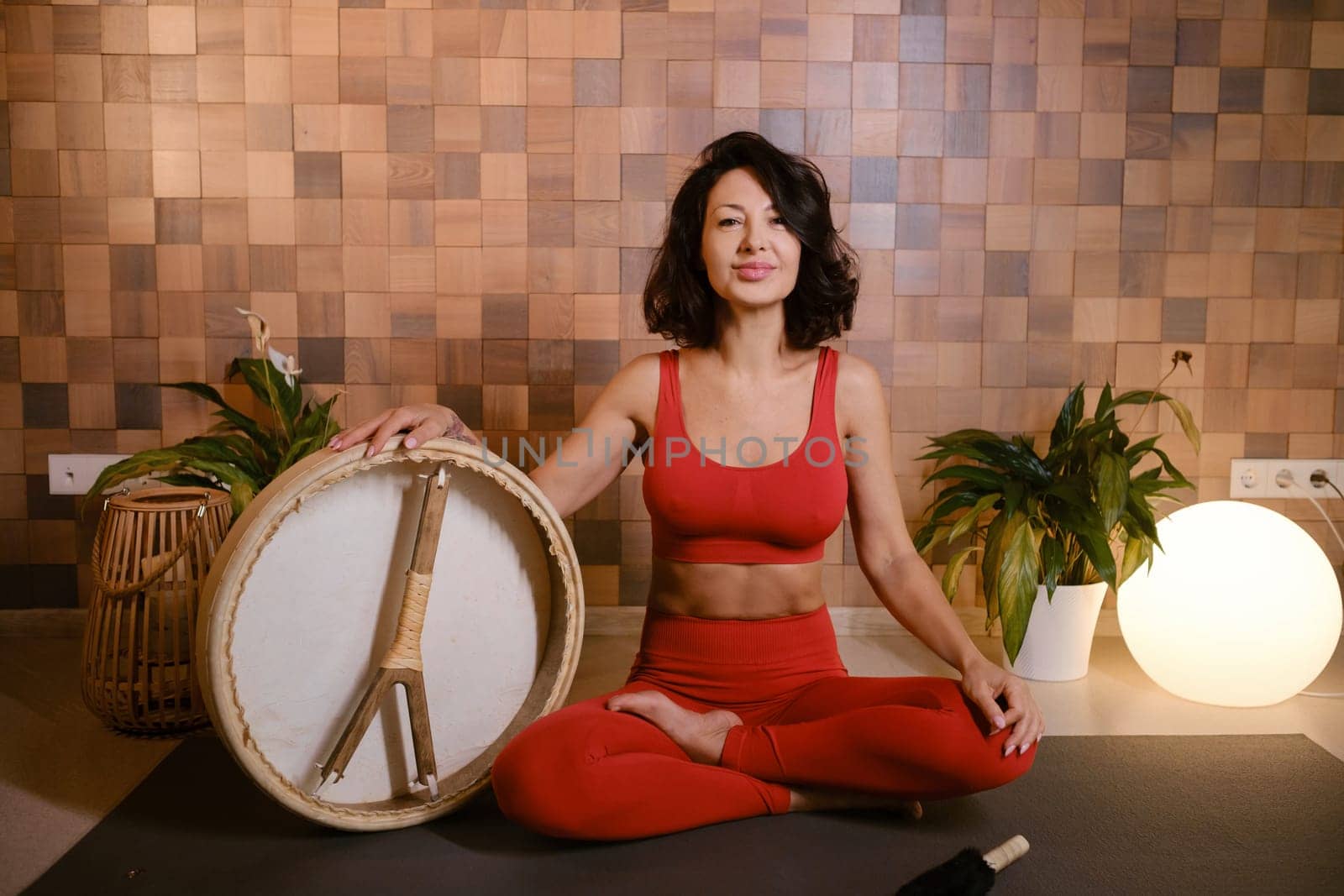 A woman in a red tracksuit sits with a drum in a gymnasium. Kalimeditation class.