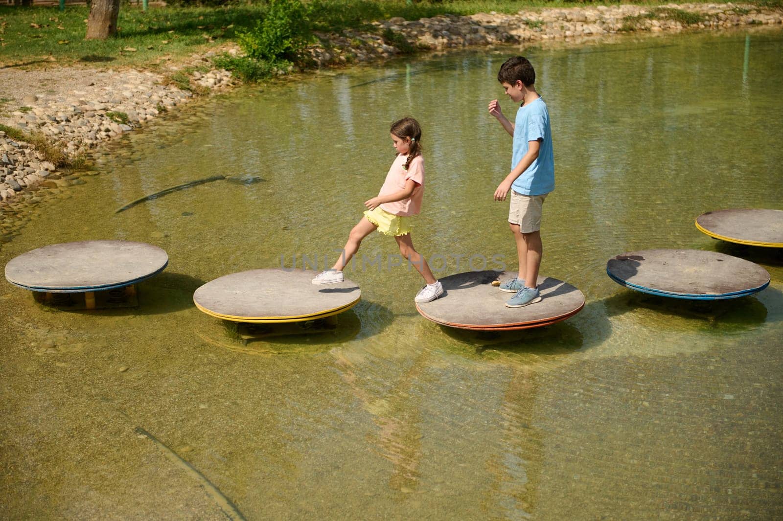 Two children balance on round stepping stones over a clear, shallow pond in a park setting.