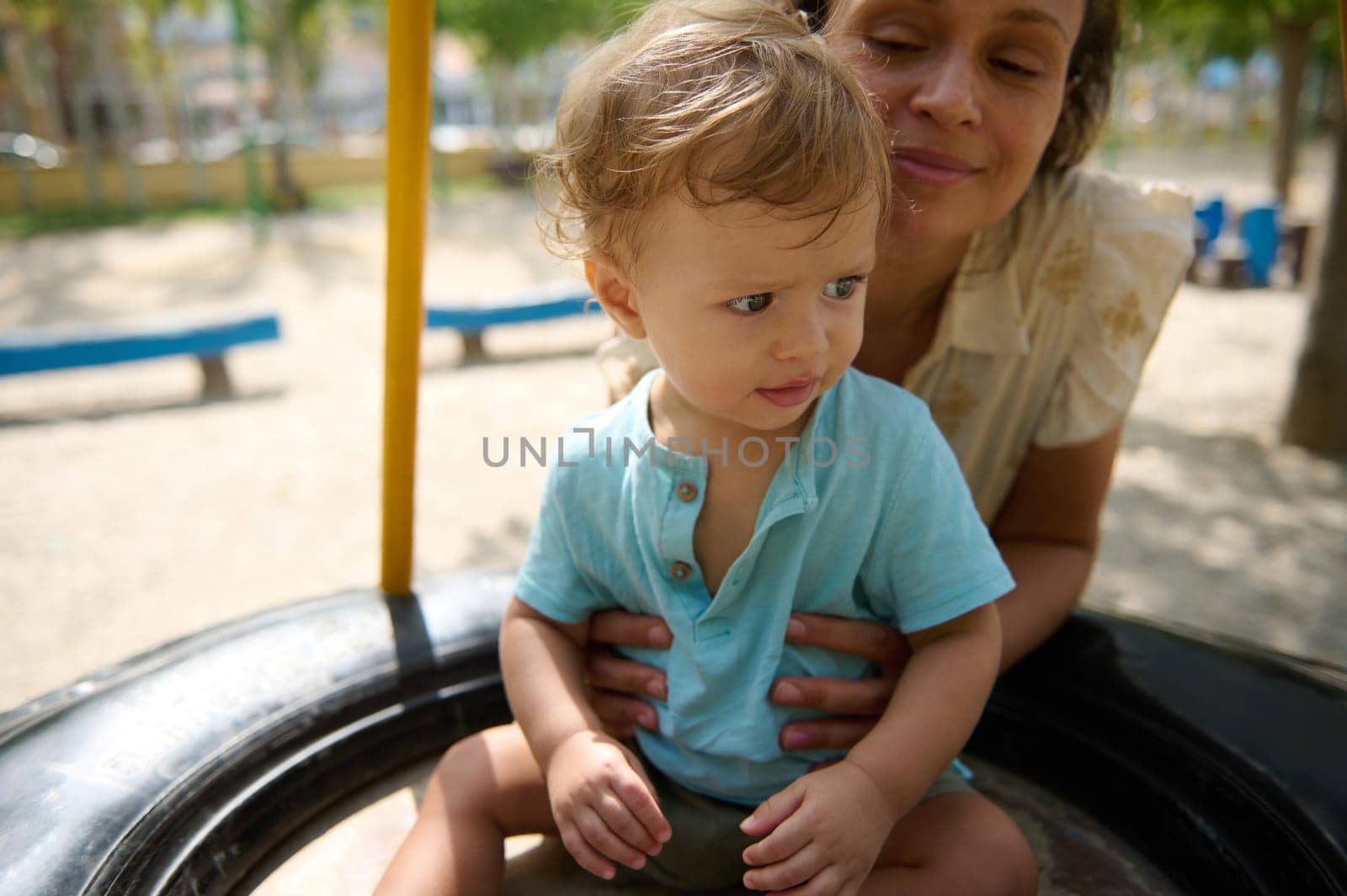 A mother lovingly holds her child on a playground tire swing on a sunny day, capturing a joyful family moment.