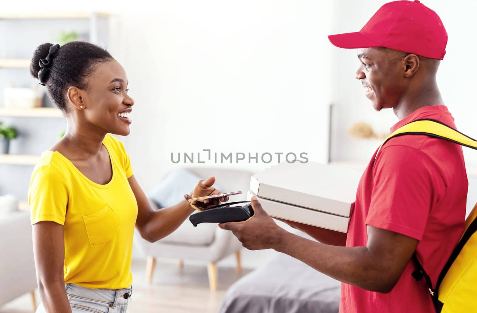 Food Delivery. African American Female Paying Courier For Pizza Putting Mobile Phone To Payment Terminal Standing Indoors. Restaurant Delivering Service Concept. Selective Focus