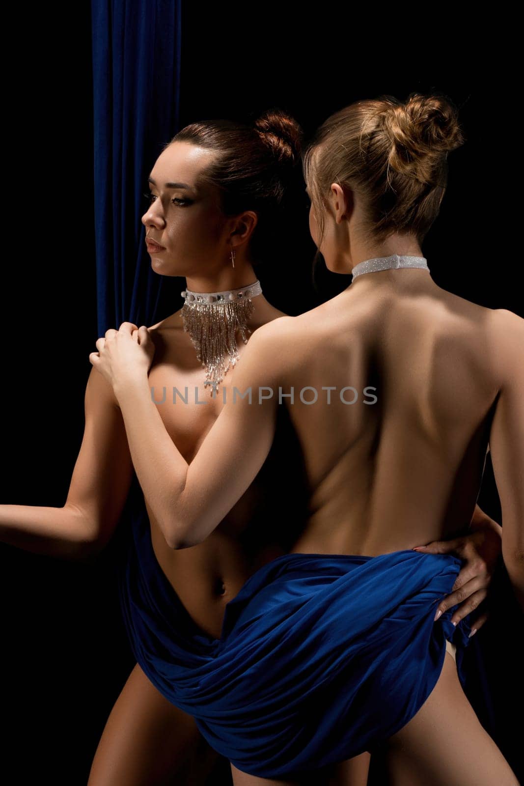 Two barefoot nude ladies wrapped in blue fabric touching each other gently while standing against black background