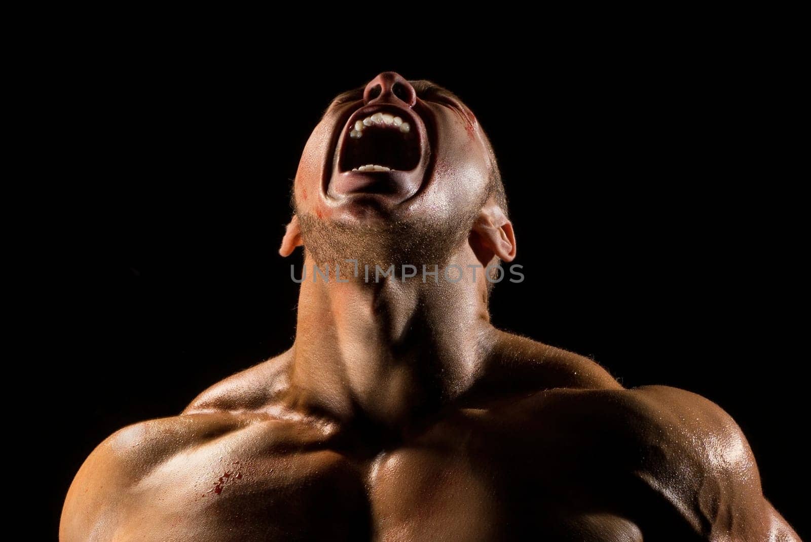 Shirtless male athlet with bandaged bloody arms screaming shot in the dark room