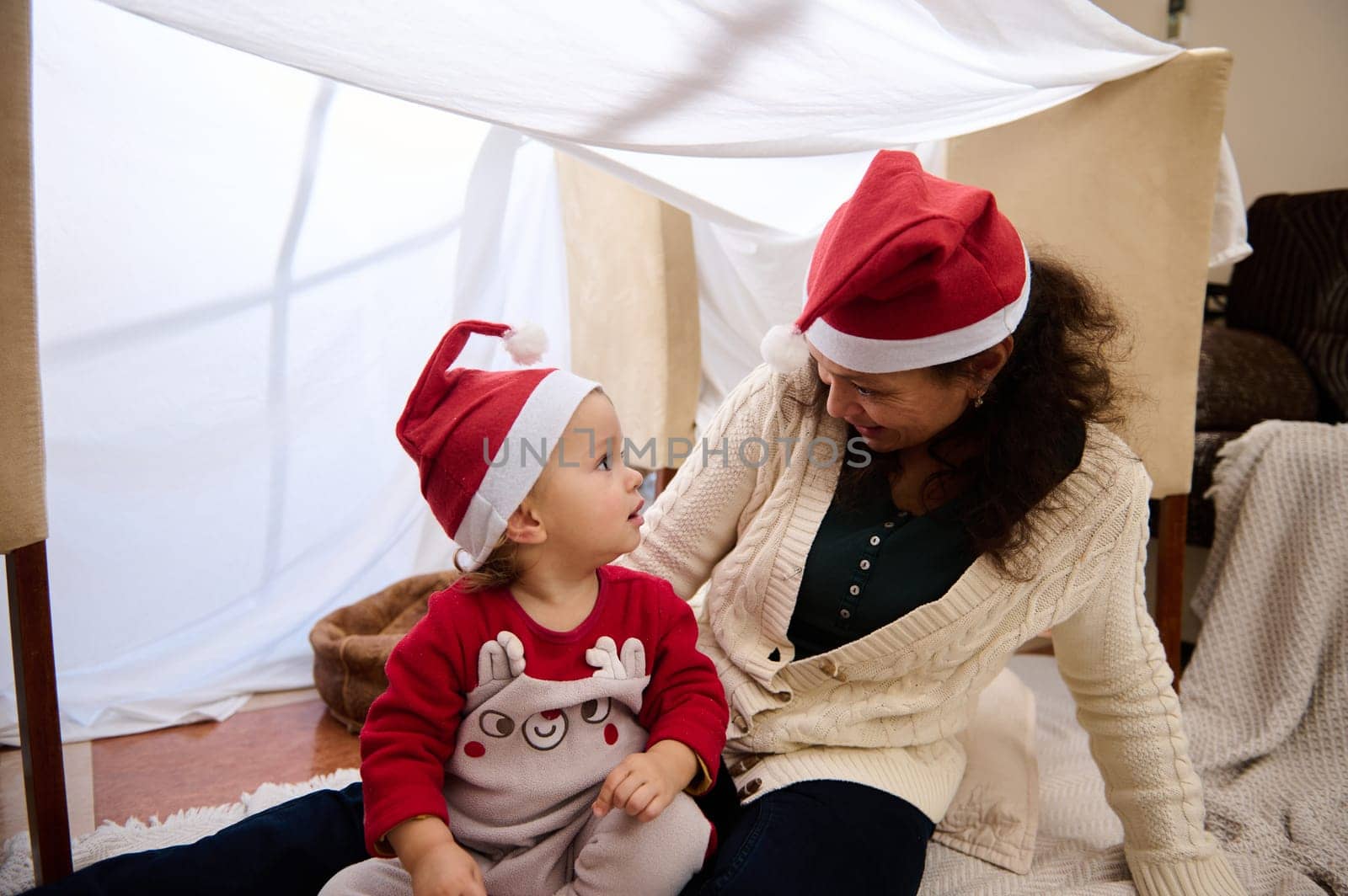 A joyful mother and her child wearing Santa hats share a playful holiday moment indoors.