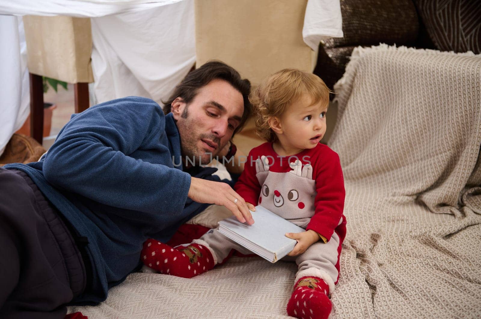 A father and his young child share a quiet moment reading a book in a homemade pillow fort, fostering a sense of warmth and family bonding.