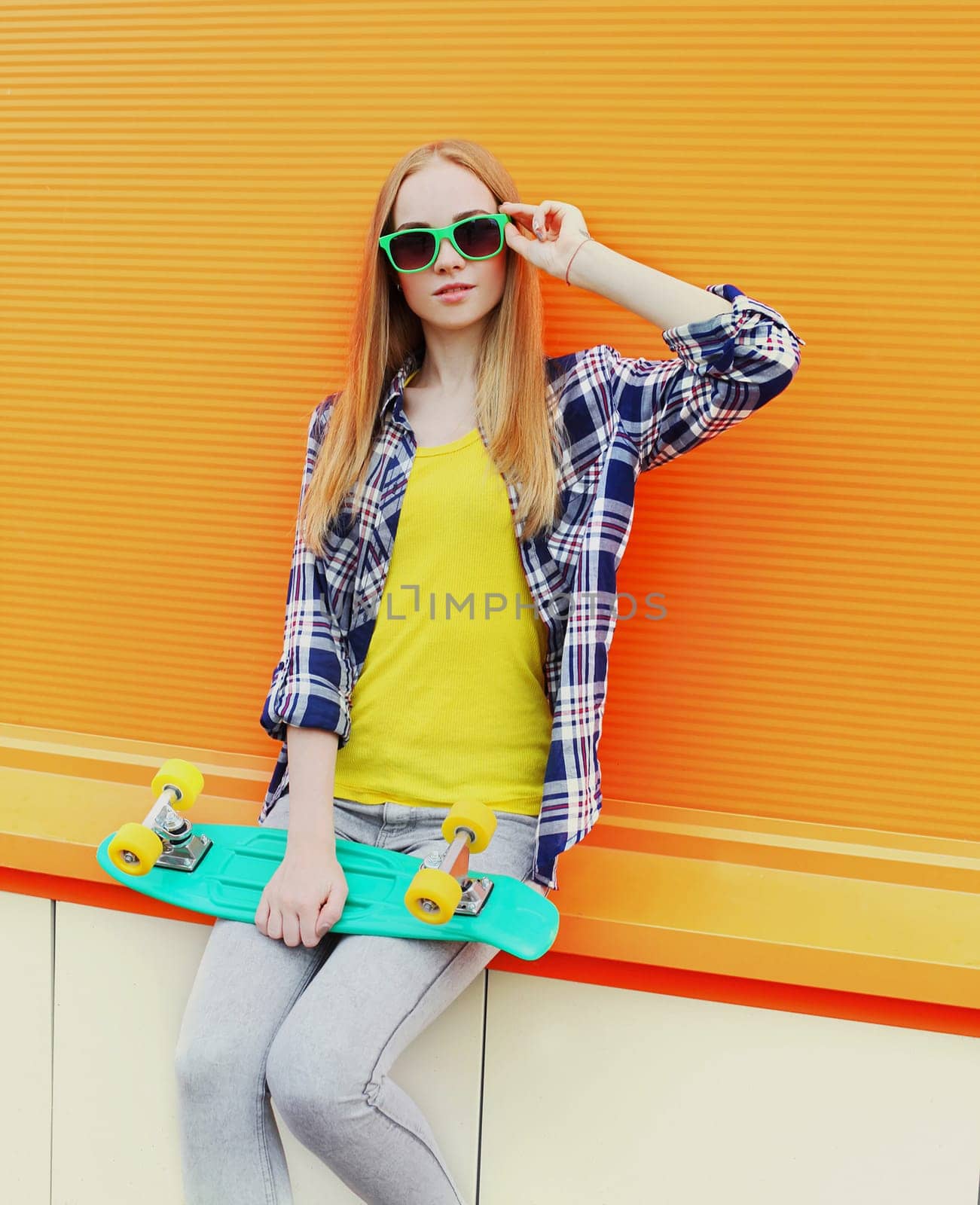 Fashion pretty blonde girl wearing a sunglasses with skateboard over colorful orange background