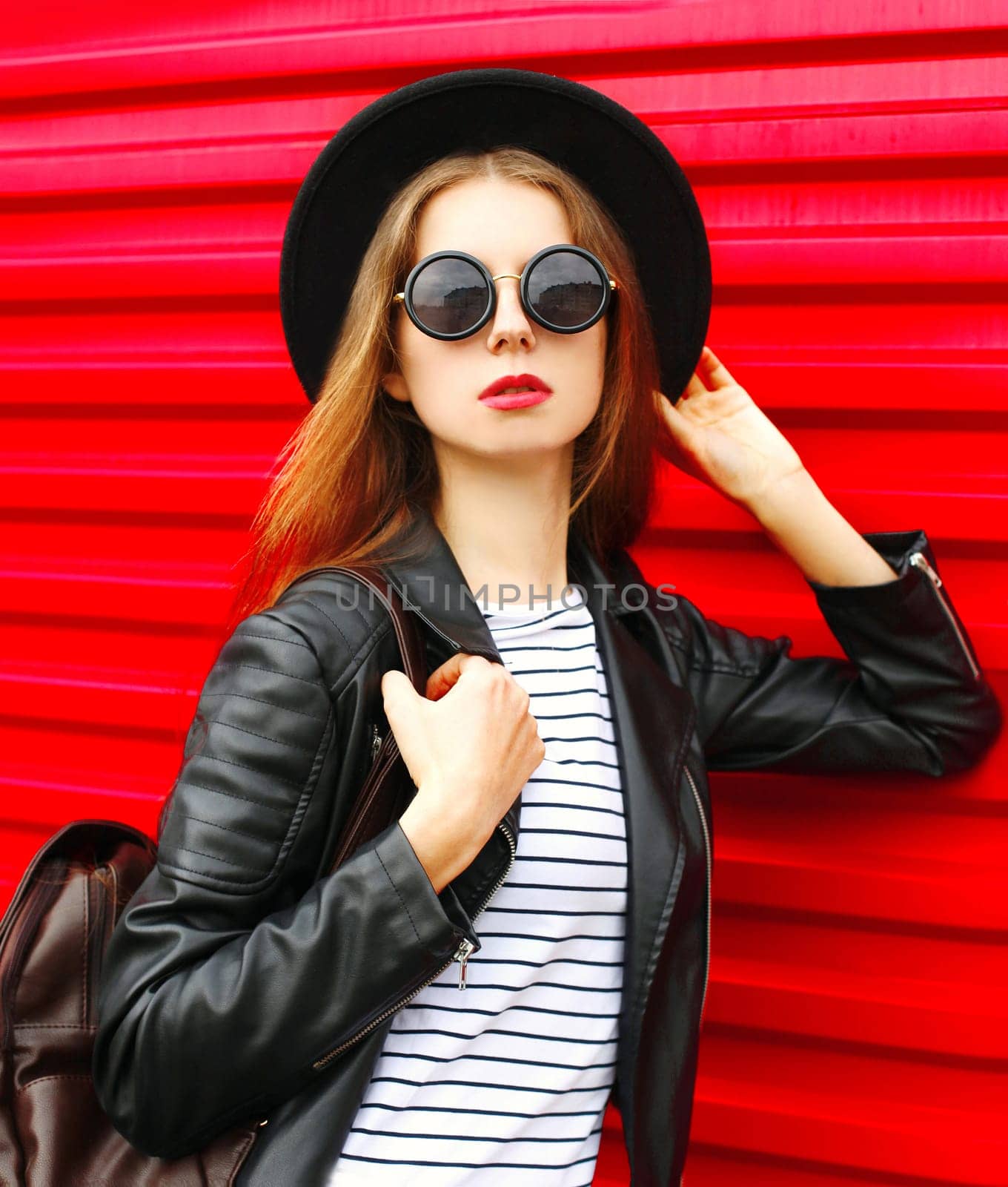 Fashion portrait young woman in black rock style hat, jacket stands with backpack on red background