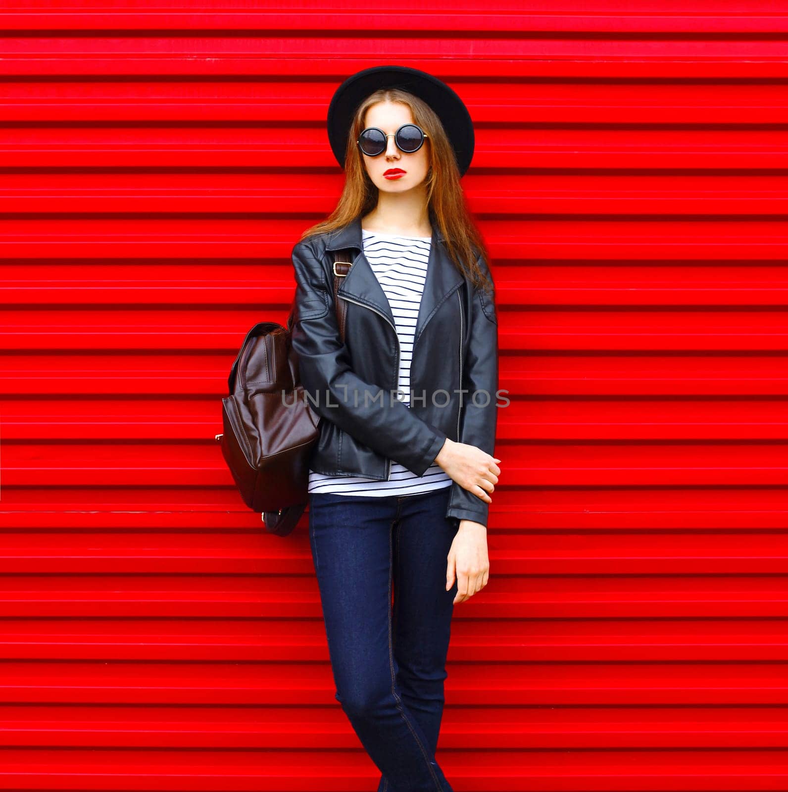 Fashion portrait young woman in black rock style hat, jacket stands with backpack on red background