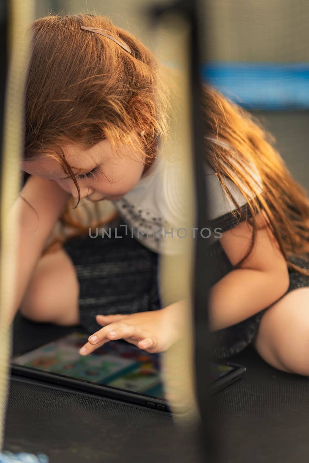 little girl sits on a trampoline and plays on a tablet