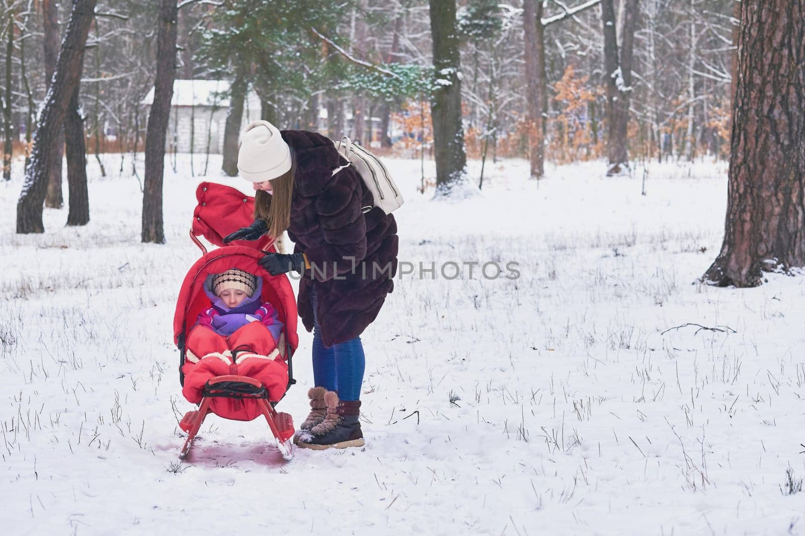 A young mother straightens the clothes of a child sitting in sled in winter park by jovani68