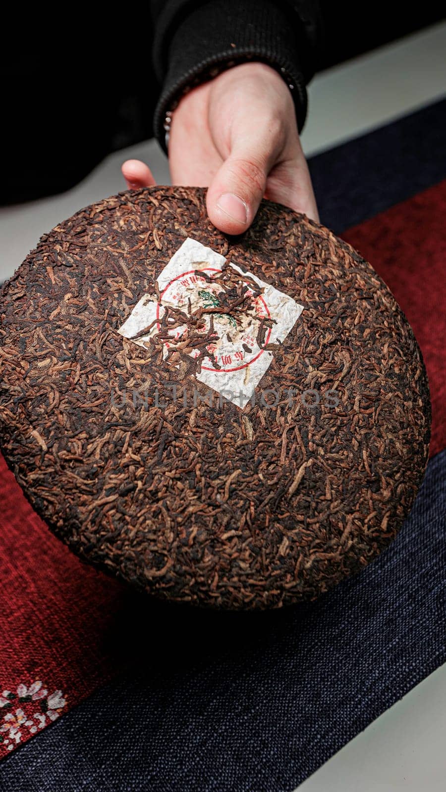 A person is holding a round compressed tea cake with intricate textures displayed beautifully on a textured surface