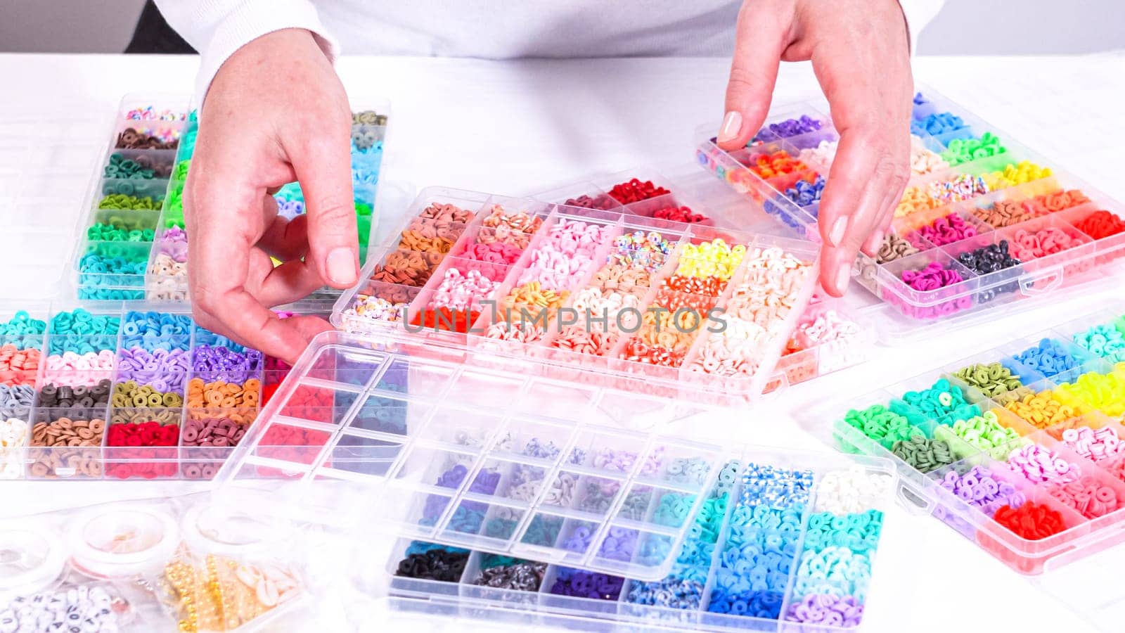 Womans hands gracefully poised over a collection of beads, sorted by color in transparent organizers. The array of beads spans a vibrant spectrum, from deep purples to bright oranges, meticulously arranged for easy selection as she embarks on creating a custom piece of jewelry.