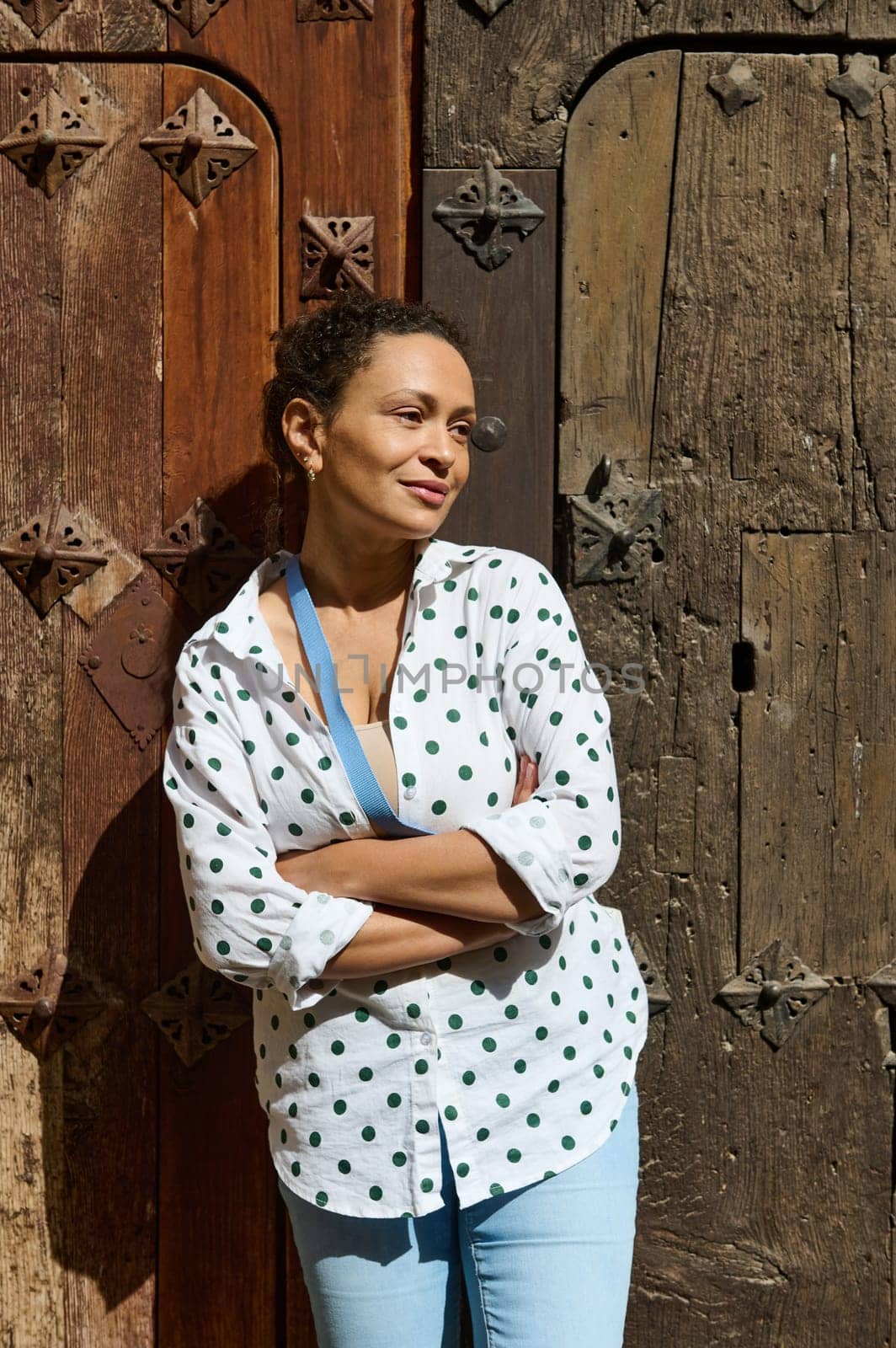 A confident woman with crossed arms stands against a rustic, intricately carved wooden door, soaking up the sunlight.