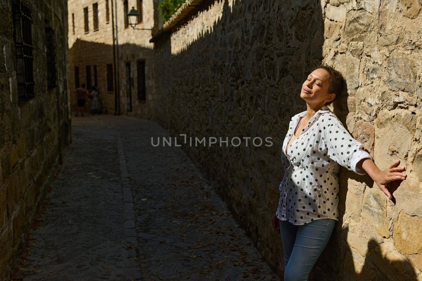 A woman in a polka dot shirt leans against a sunlit stone wall, savoring the tranquility of a historic cobblestone alley.