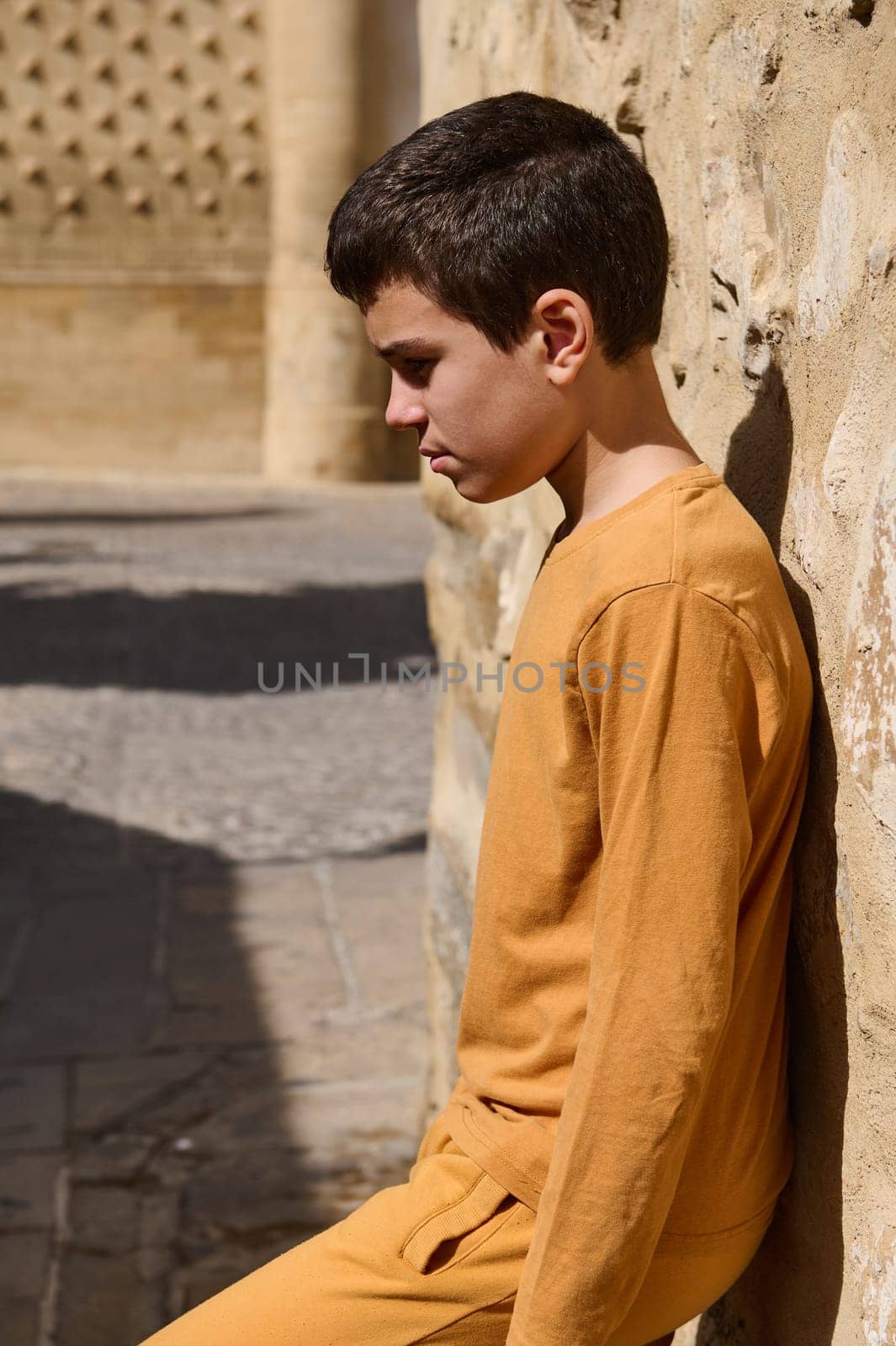 A thoughtful young boy in mustard attire leans against a weathered stone wall under sunlight, immersed in thought.
