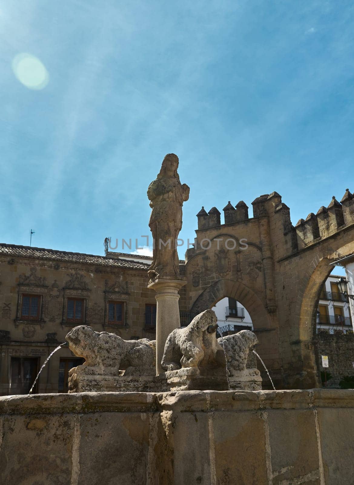 A historic stone fountain adorned with statues, set against a picturesque blue sky. The fountain features intricate details and reflects the charm of ancient architecture.