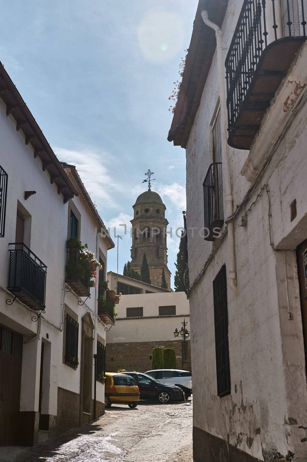 A picturesque, narrow alleyway with old buildings and a church tower in the background, under a clear blue sky.