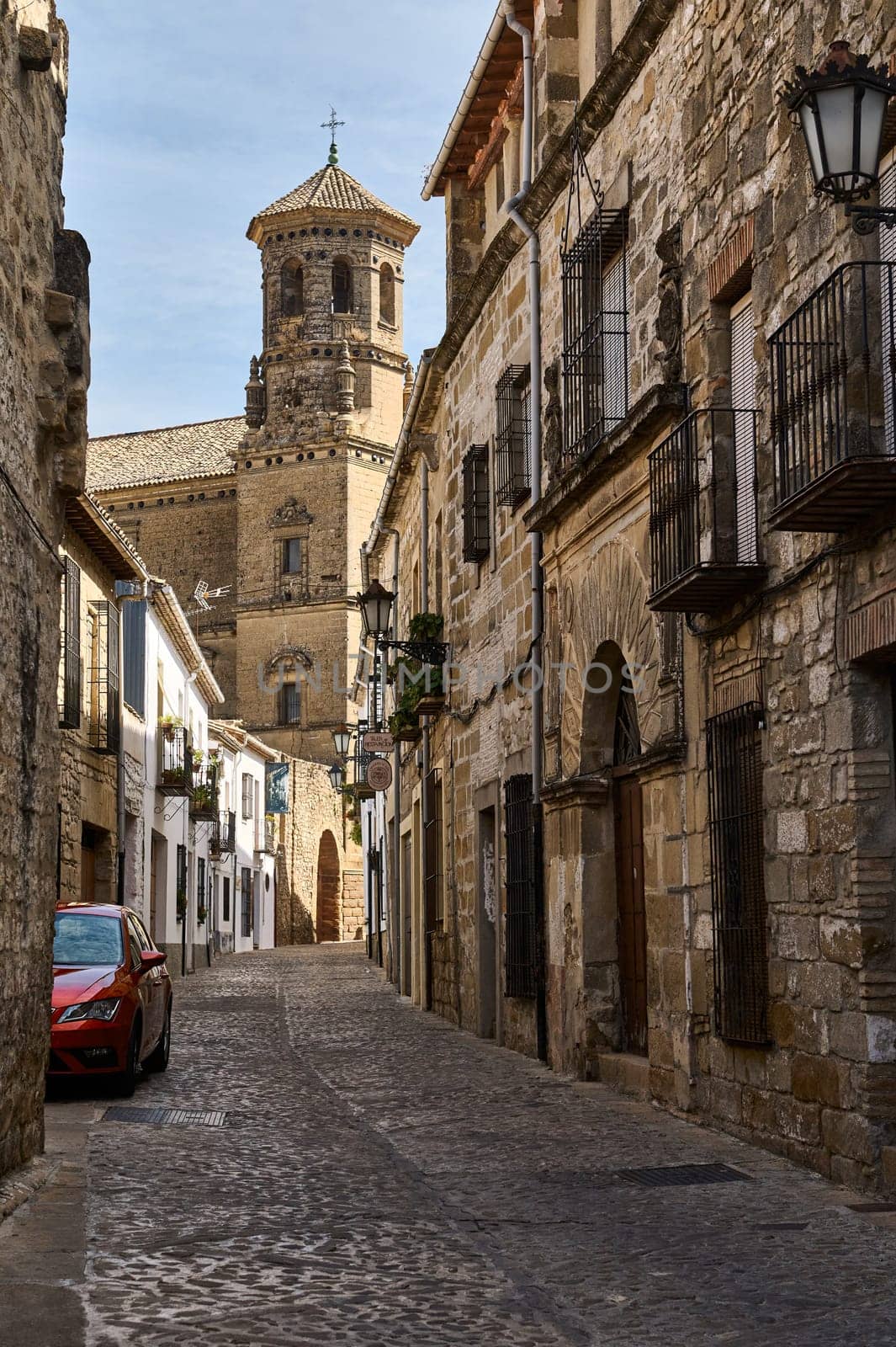 A charming European street featuring cobblestone paths, traditional stone facades, and a prominent historic church tower under a clear sky.