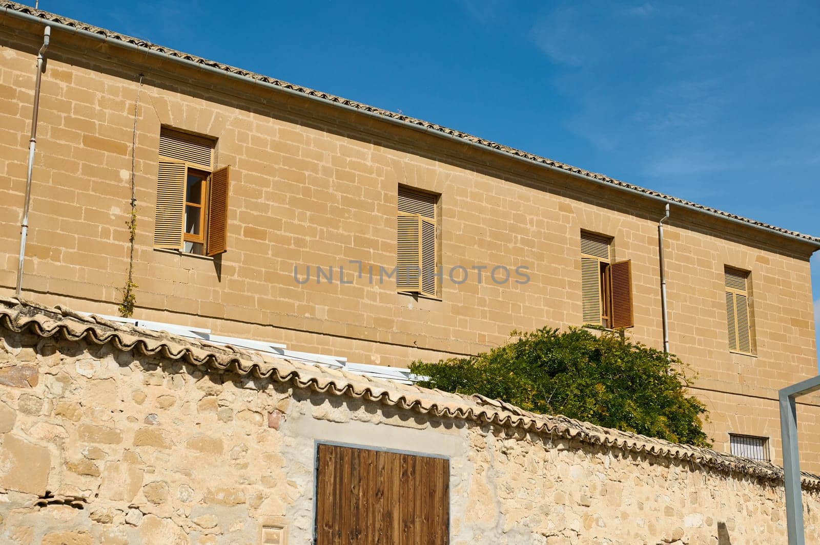 A charming historic stone building with open wooden shutters, basking in the sunlight against a clear blue sky, showcasing classic architecture.