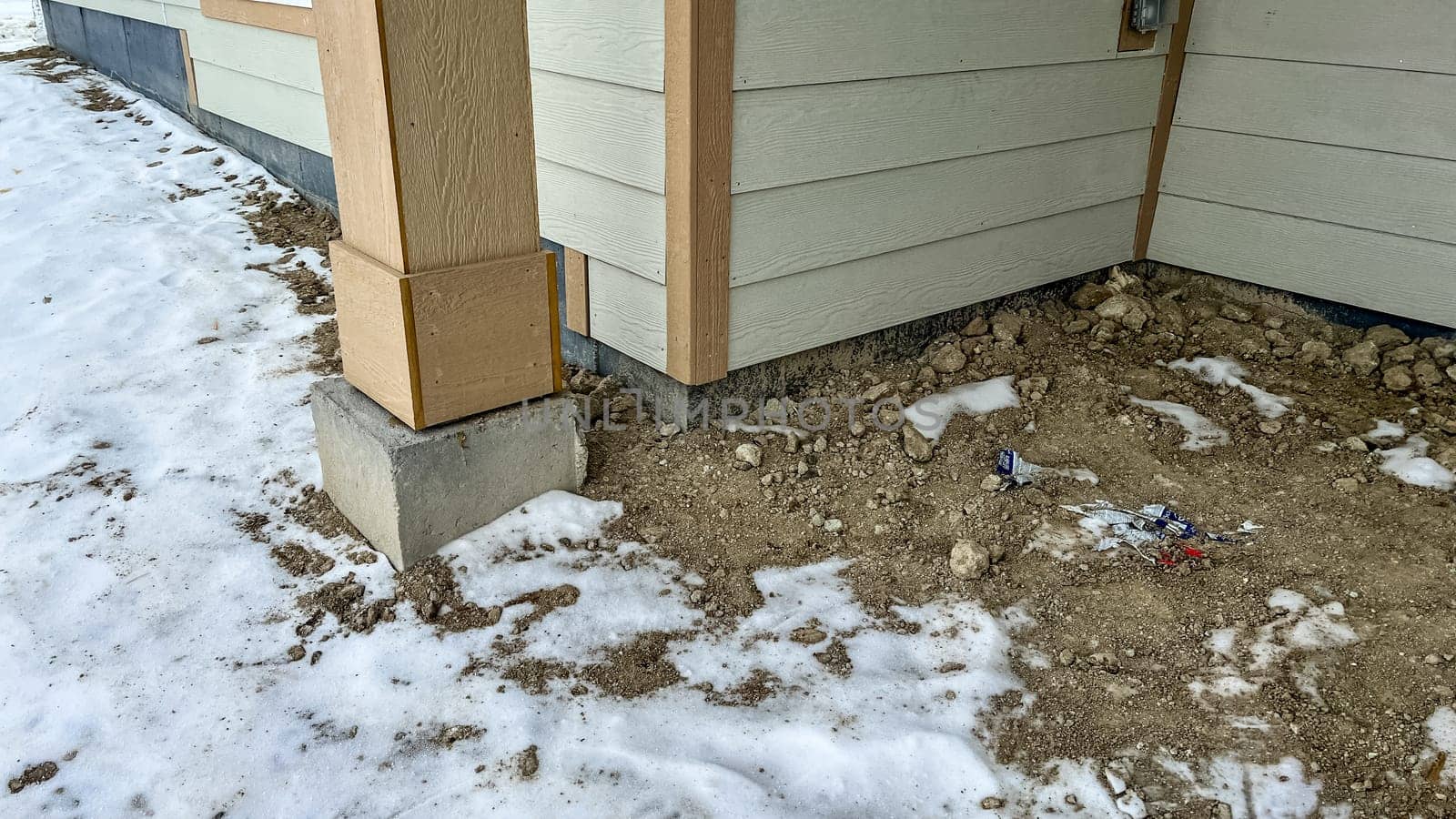Close-up view of a house foundation surrounded by dirt and light patches of melting snow. Wooden posts and siding are visible, suggesting construction or a backyard area in winter conditions.