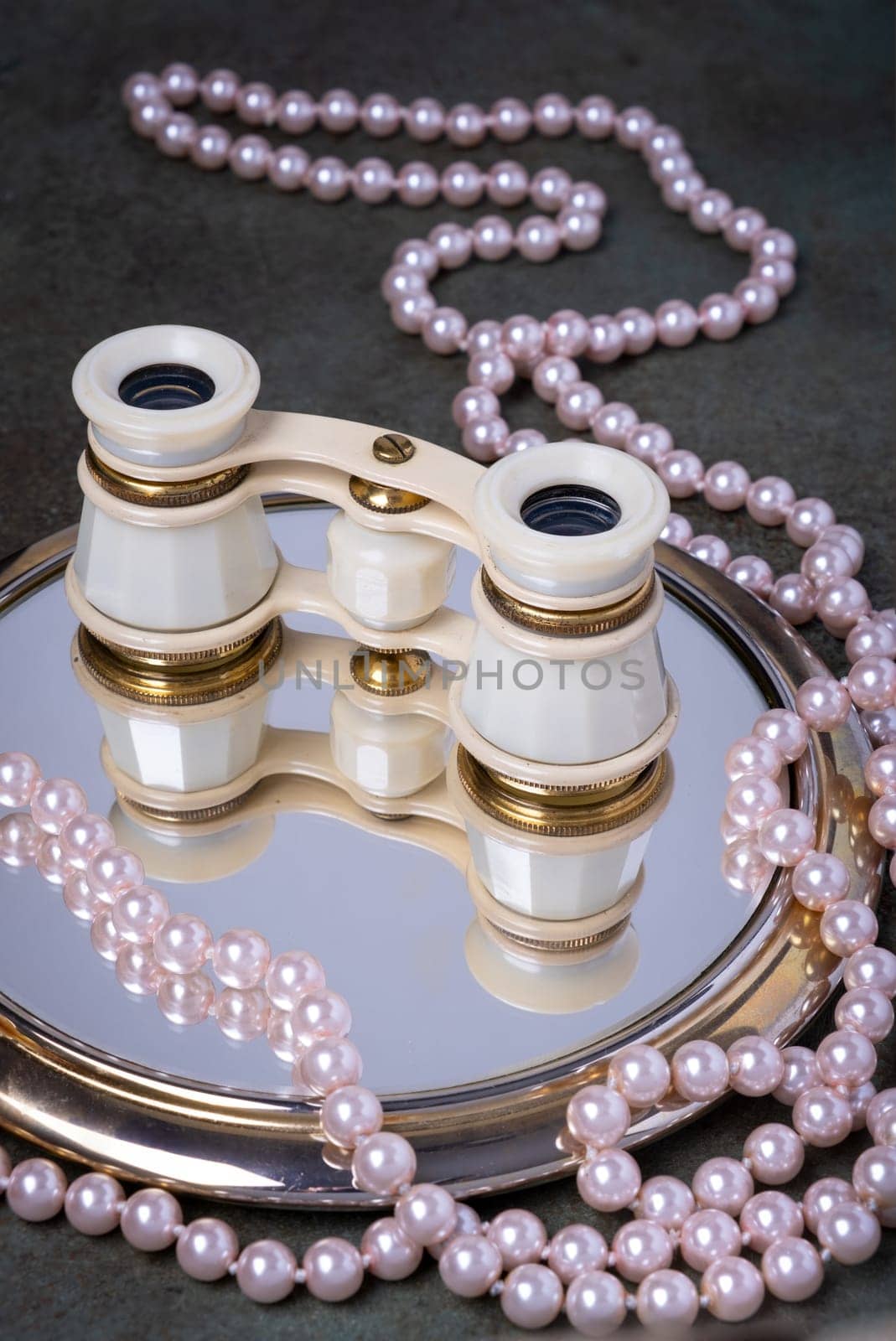 Binoculars rest on mirror surrounded by pearl necklaces. The image evokes elegance and timelessness.