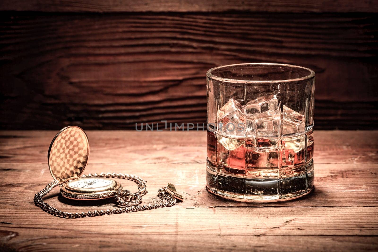 A retro style image of a glass of liquor with ice and a pocket watch on wood.