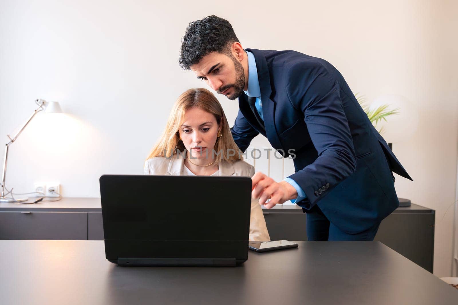 Professional man and woman working together on a laptop in a modern office. The woman points at the screen, explaining details while the man listens attentively.