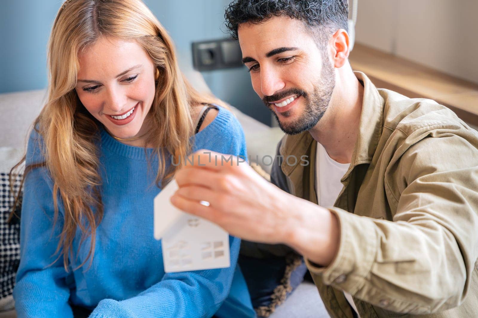 A cheerful couple sits in a cozy home, holding a small house model. Their smiles and relaxed posture create a warm atmosphere, perfect for themes of home ownership, planning, and relationships.