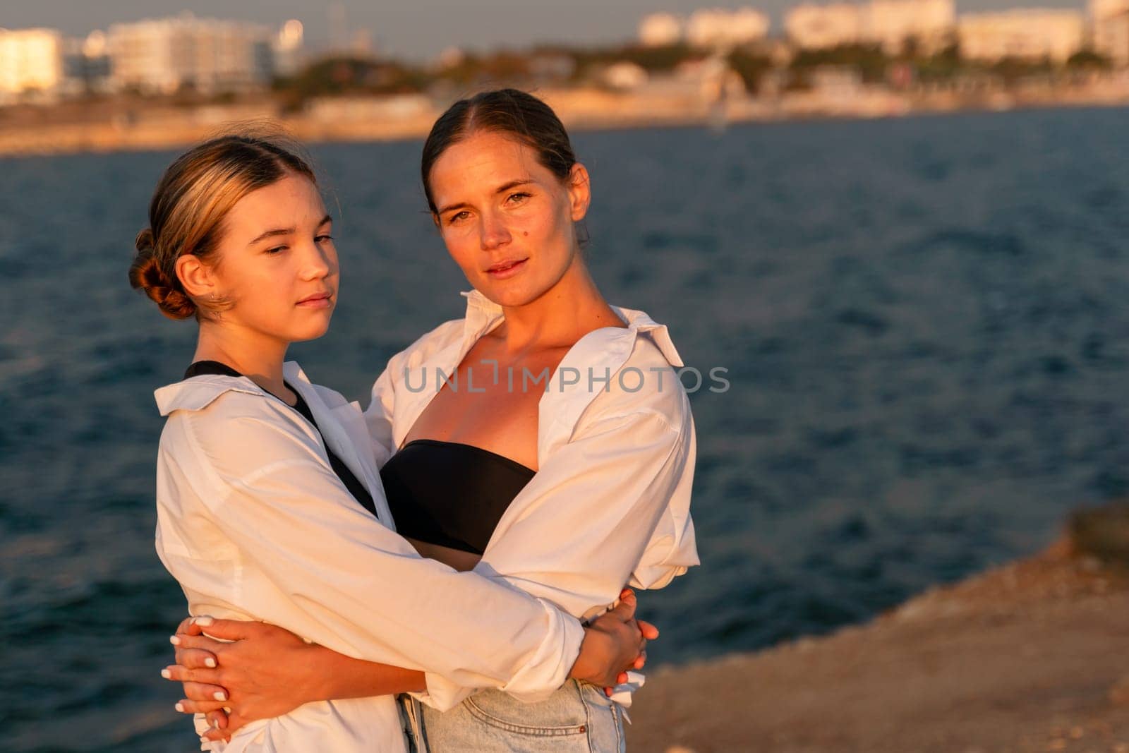 Two women, one in a white shirt and the other in a black bandeau top and white shirt, embrace on a rocky shore at sunset. The sea and a distant city are visible in the background.