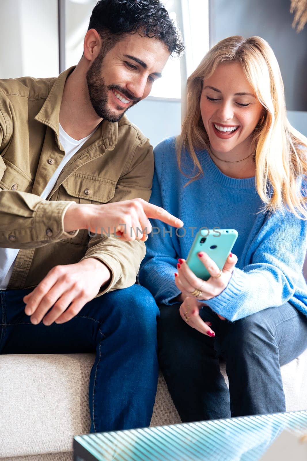 Young man and woman sitting on a couch, sharing a moment of excitement while looking at a smartphone. Casual outfits and a relaxed indoor environment.
