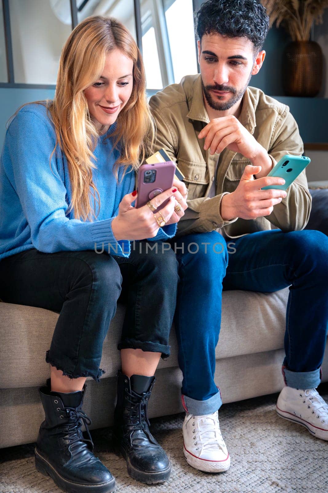 Young man and woman sitting on a couch, sharing a moment of excitement while looking at a smartphone. Casual outfits and a relaxed indoor environment.