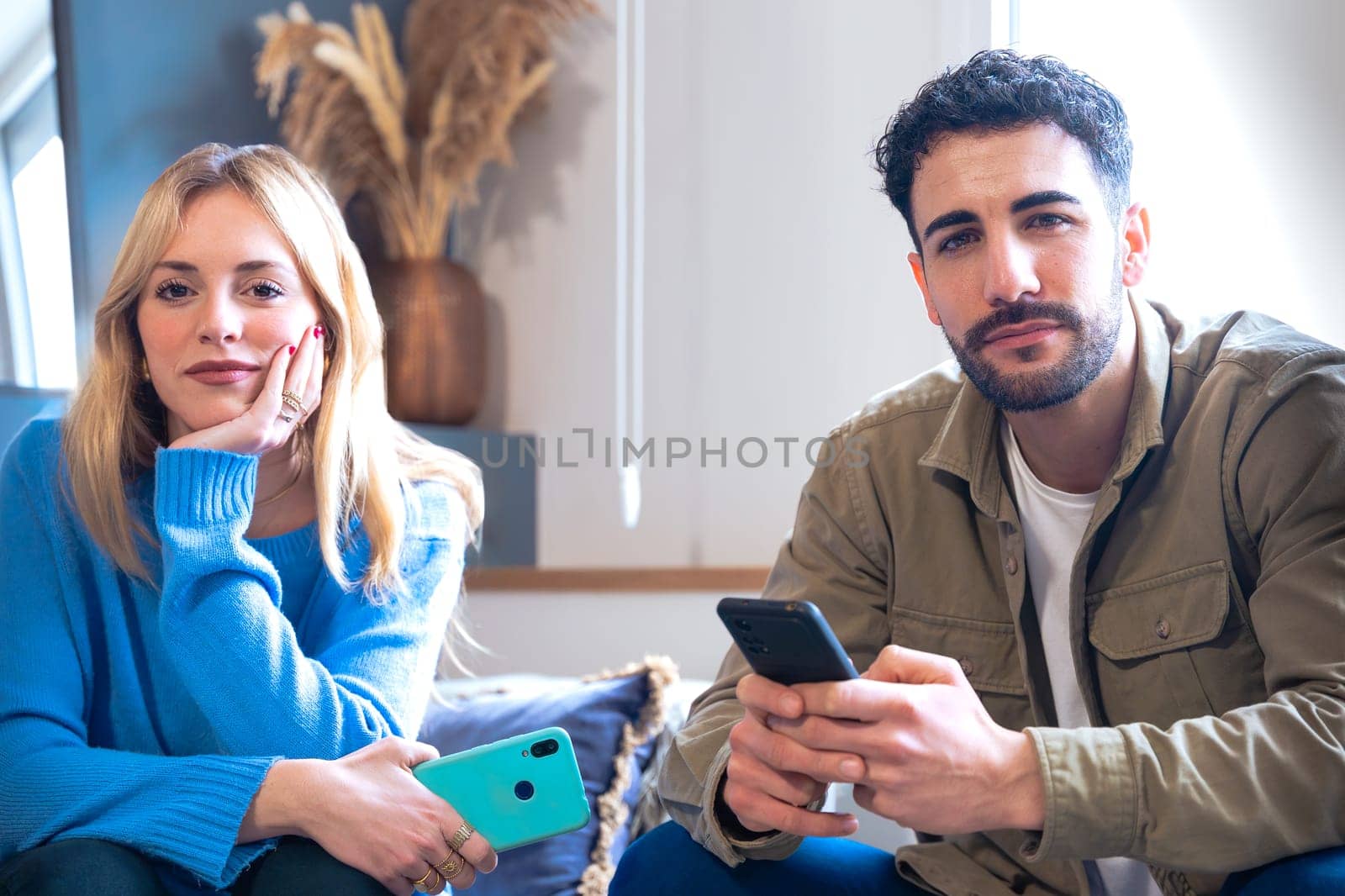 A young couple sitting indoors using smartphones, expressing thoughtful emotions. The woman holds her phone with a pensive look, while the man glances with a neutral expression