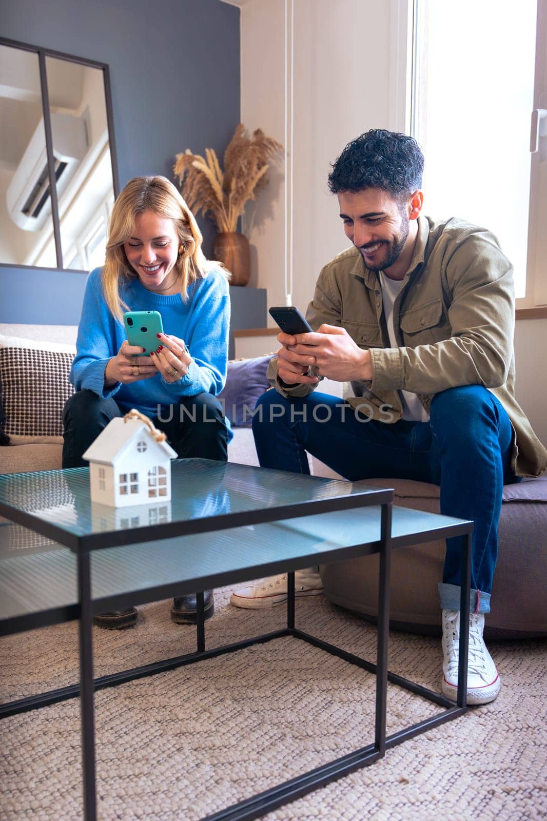 A young couple seated in a modern living room, laughing while using their smartphones. A miniature house decorates the glass coffee table, enhancing the cozy atmosphere.Vertical