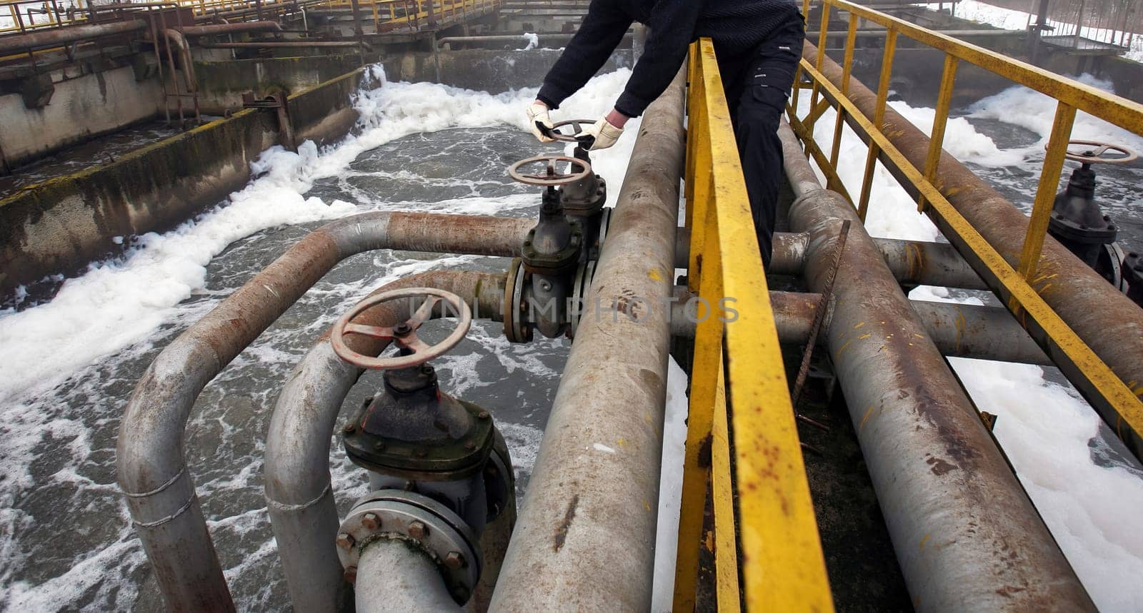 A worker adjusts valves on large pipes at an old water treatment facility while water flows through the system, highlighting maintenance efforts.