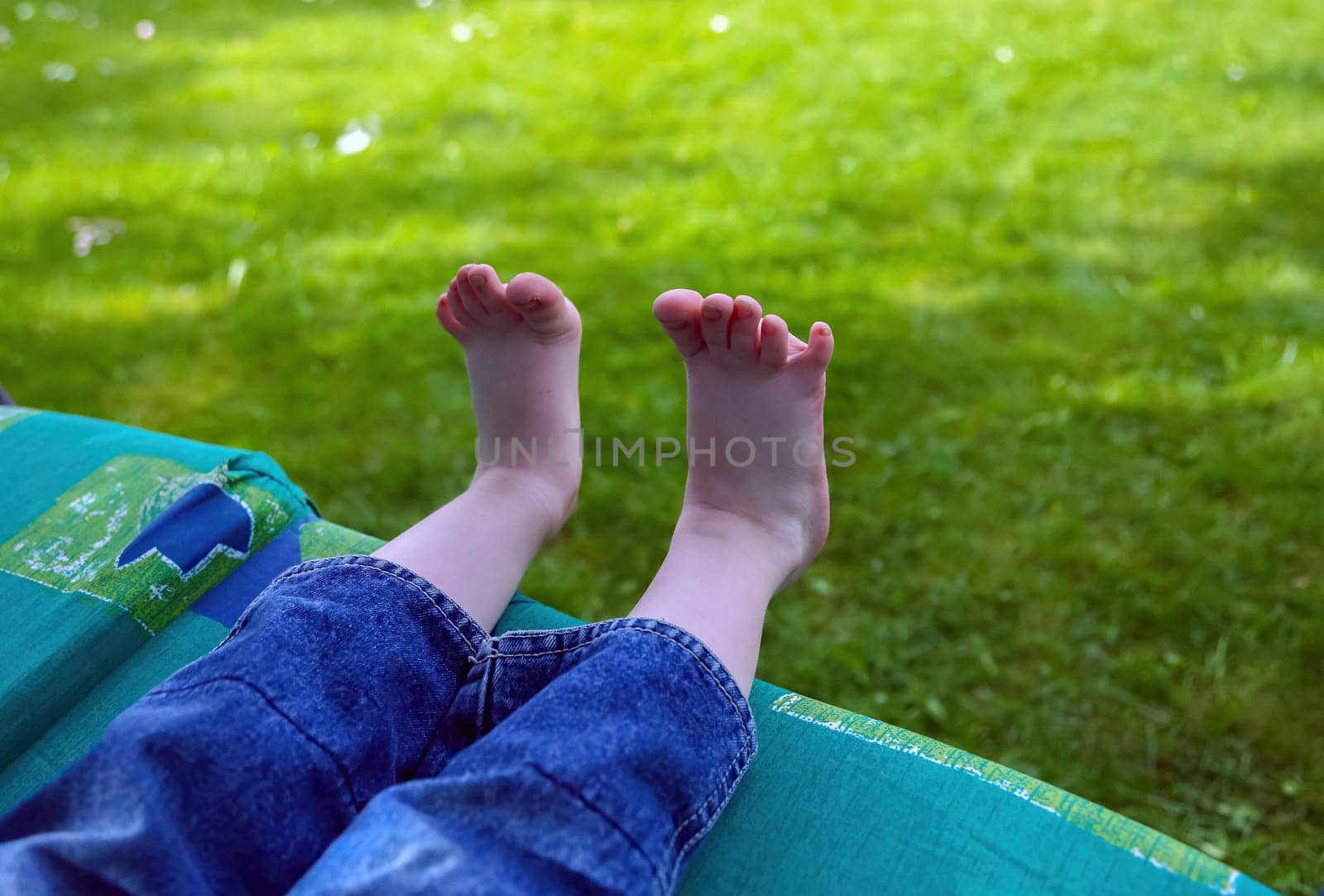 A child's bare feet dangle from a blanket, enjoying a sunny afternoon in a lush green garden, evoking feelings of relaxation.