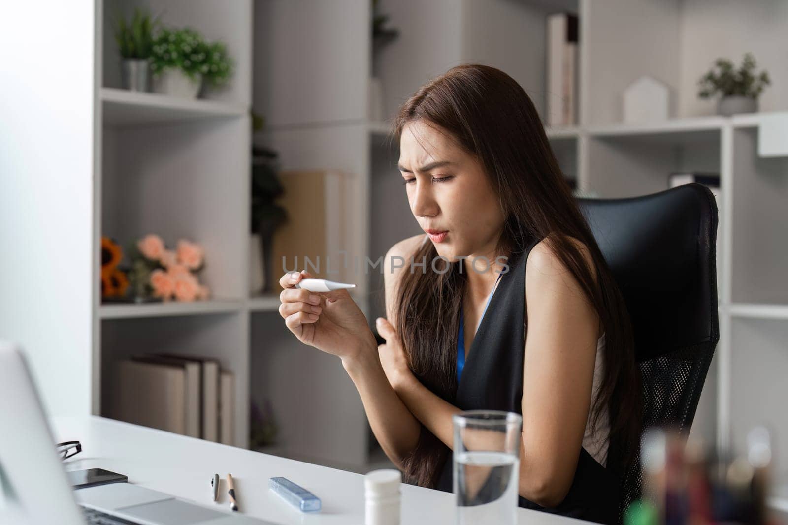 A young female office worker sits at her desk, visibly unwell with cold symptoms, in a modern office environment.