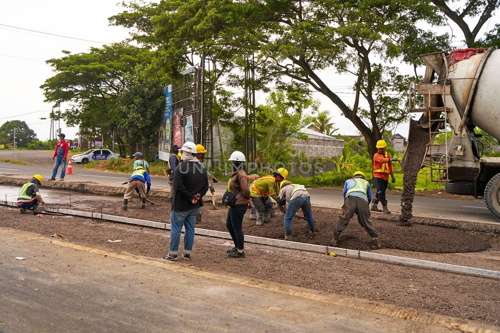 Road repairs on a major highway in Asia. Uniformed workers wearing protective helmets pour asphalt near a concrete mixing machine. Bali, Indonesia - 12.16.2022