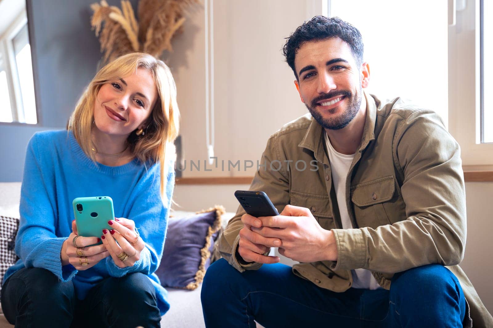 A cheerful young couple sitting indoors, both holding smartphones and smiling warmly. The woman leans slightly, creating a relaxed and friendly atmosphere in a cozy setting.
