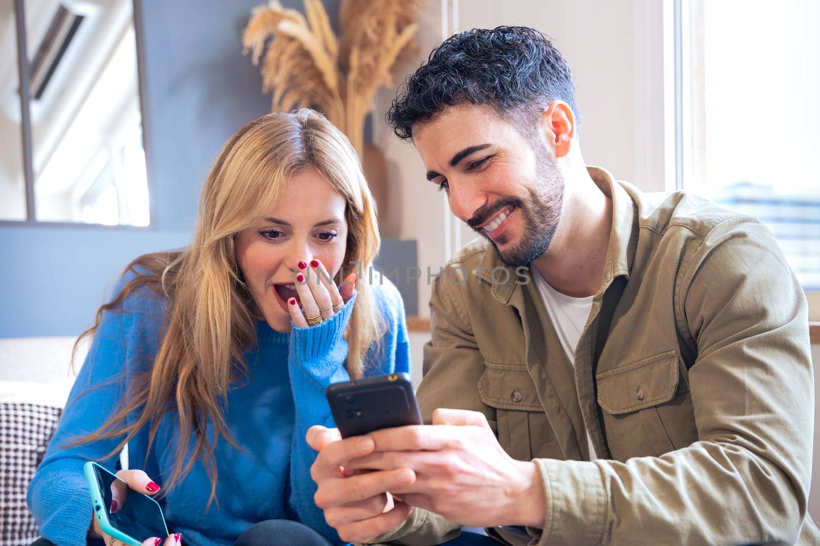 A young couple seated indoors sharing a surprising and joyful moment while looking at a smartphone. The woman covers her mouth in amazement as the man smiles warmly.