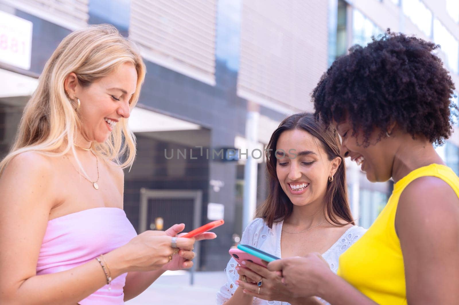 Three cheerful young women gathers around a red smartphone, reacting with excitement. They are dressed casually, standing outdoors in a modern urban environment, enjoying a fun moment together.