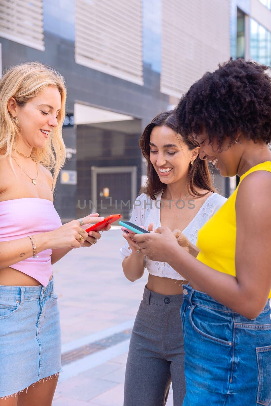 Three cheerful young women gathers around a red smartphone, reacting with excitement. They are dressed casually, standing outdoors in a modern urban environment, enjoying a fun moment together.