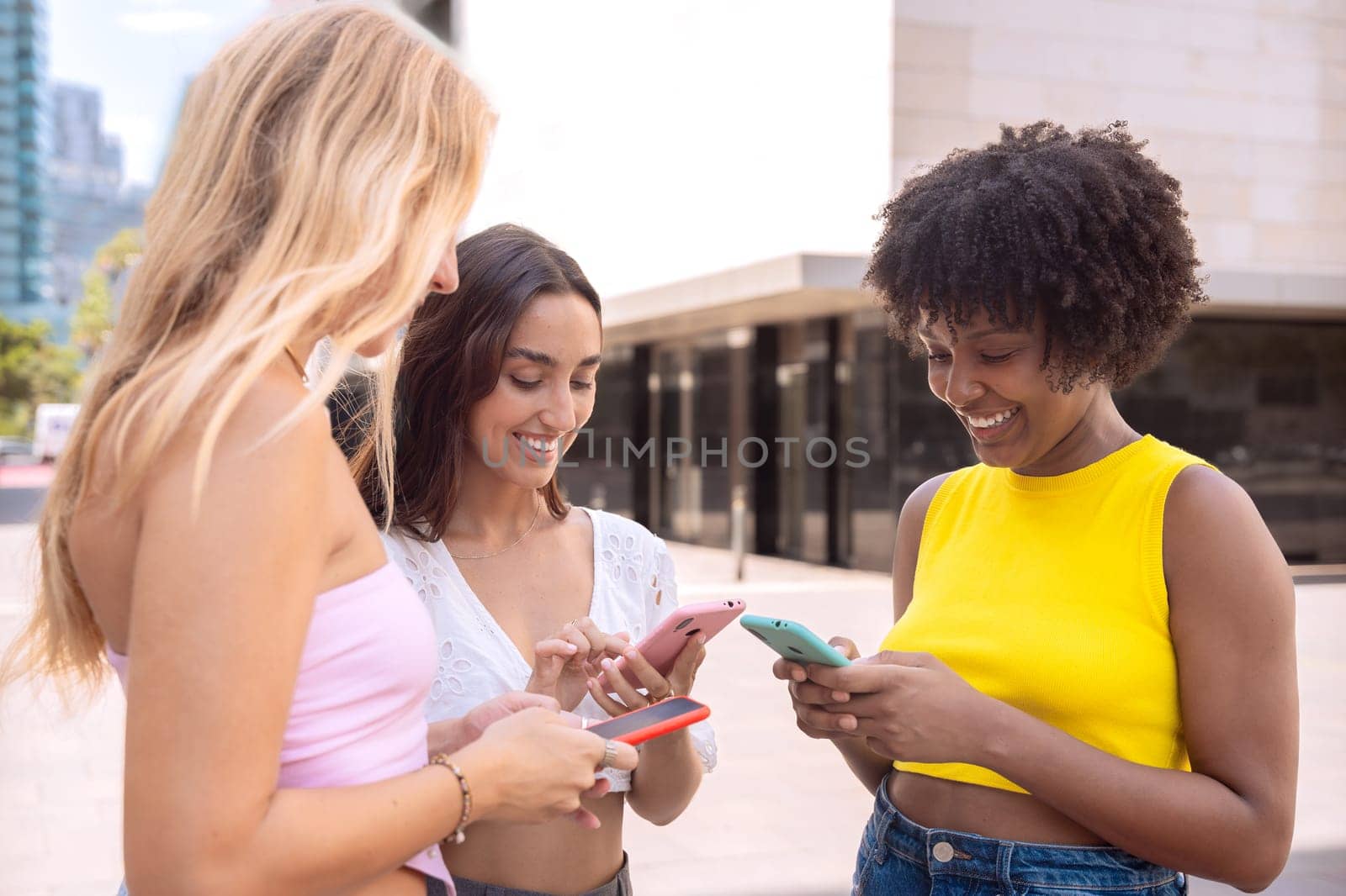 Three cheerful young women gathers around a red smartphone, reacting with excitement. They are dressed casually, standing outdoors in a modern urban environment, enjoying a fun moment together.